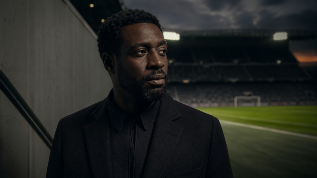 Portrait éditorial d’un homme africain pensif au bord d’un stade de football, dans une ambiance cinématographique sombre.