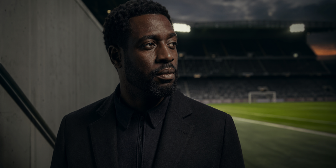 Portrait éditorial d’un homme africain pensif au bord d’un stade de football, dans une ambiance cinématographique sombre.