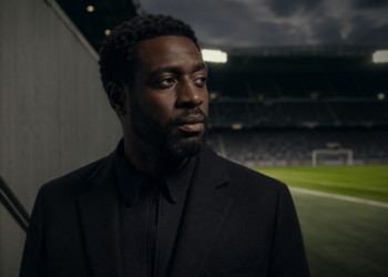 Portrait éditorial d’un homme africain pensif au bord d’un stade de football, dans une ambiance cinématographique sombre.