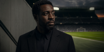 Portrait éditorial d’un homme africain pensif au bord d’un stade de football, dans une ambiance cinématographique sombre.